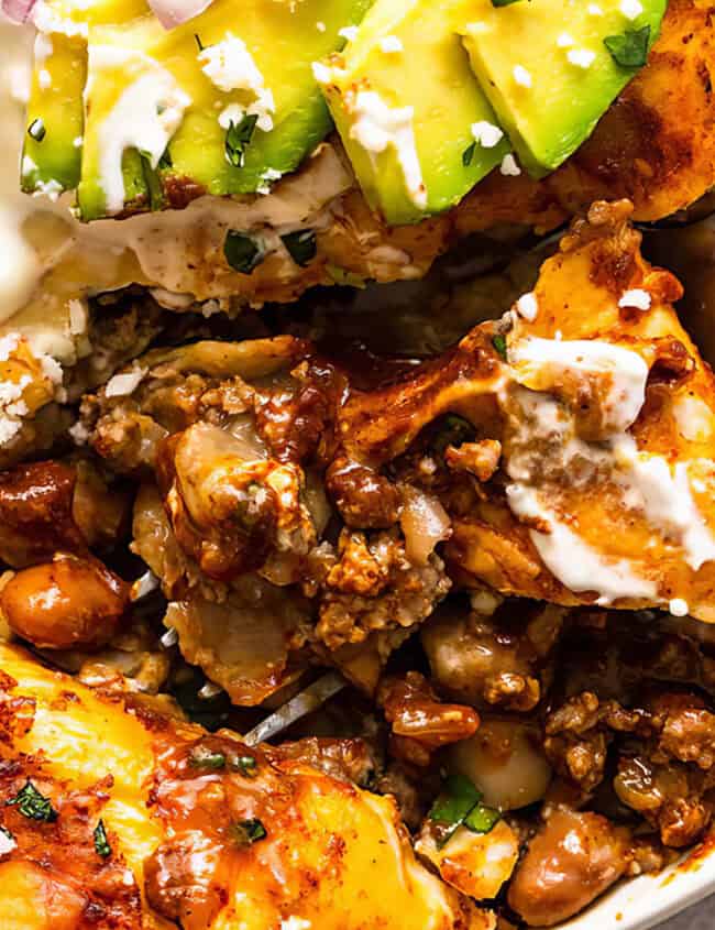 A close-up of a plate of enchiladas topped with cheese, sour cream, diced red onions, and avocado slices. The enchiladas are garnished with chopped cilantro and are filled with a savory mixture of beans and meat—perfect for lovers of ground beef recipes. A fork is placed beside the food.