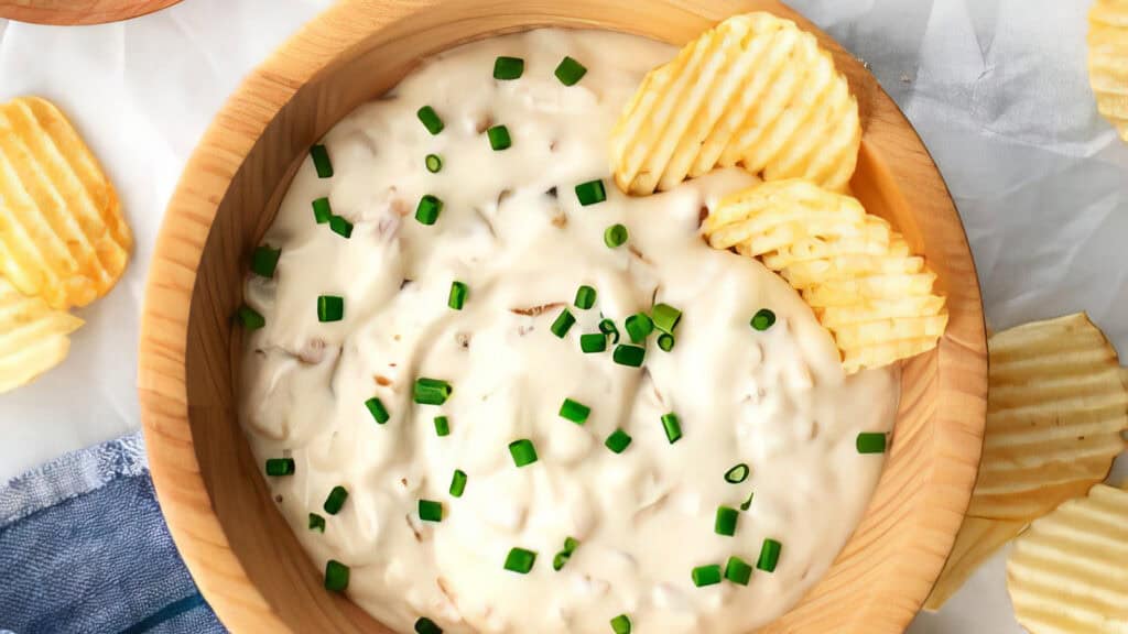 A wooden bowl filled with creamy dip garnished with chopped chives, accompanied by ridged potato chips resting on the edge. Scattered chips and a blue cloth napkin are visible around the bowl on a light surface – a perfect setup for exploring new dip recipes.