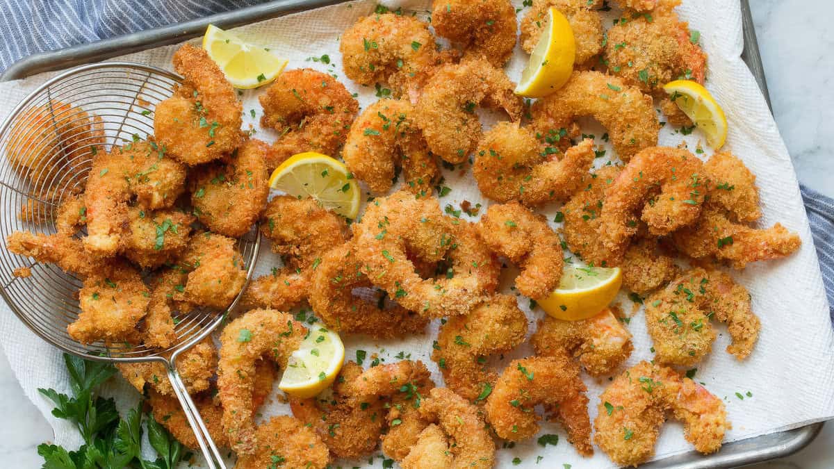 A tray filled with golden and crispy fried shrimp garnished with fresh parsley and lemon wedges. Some shrimp sit atop a wire skimmer on the left side of the tray, and a striped cloth is partially visible to the upper right—a breaded recipe delight.