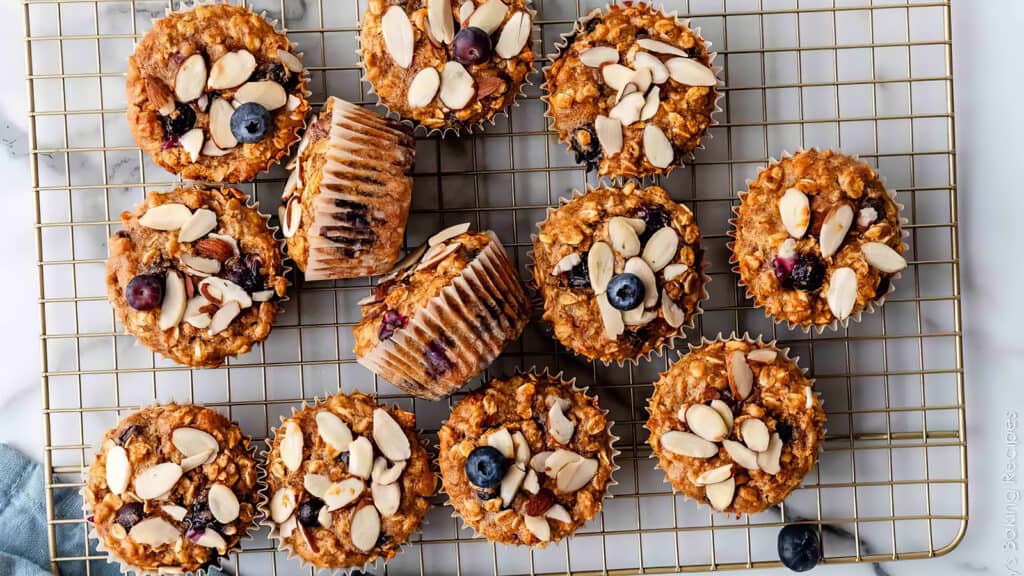 A dozen freshly baked muffins topped with almond slices and blueberries are arranged on a cooling rack. One muffin is tilted on its side, showcasing its texture and paper liner, inviting you to explore various muffin recipes. The background features a light-colored surface, hinting at a casual kitchen setting.