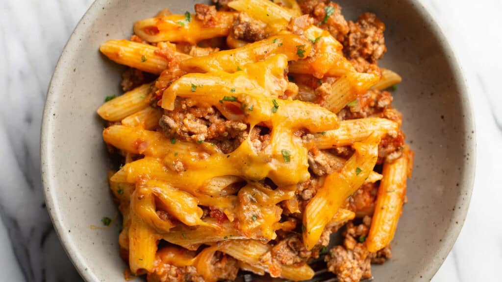 A bowl of cheesy pasta mixed with ground beef and garnished with fresh herbs. The pasta appears to be penne, and it is covered in a generous layer of melted yellow cheese, creating a comforting and hearty dish. Perfect for those seeking delicious ground beef recipes, the bowl rests on a light-colored surface.