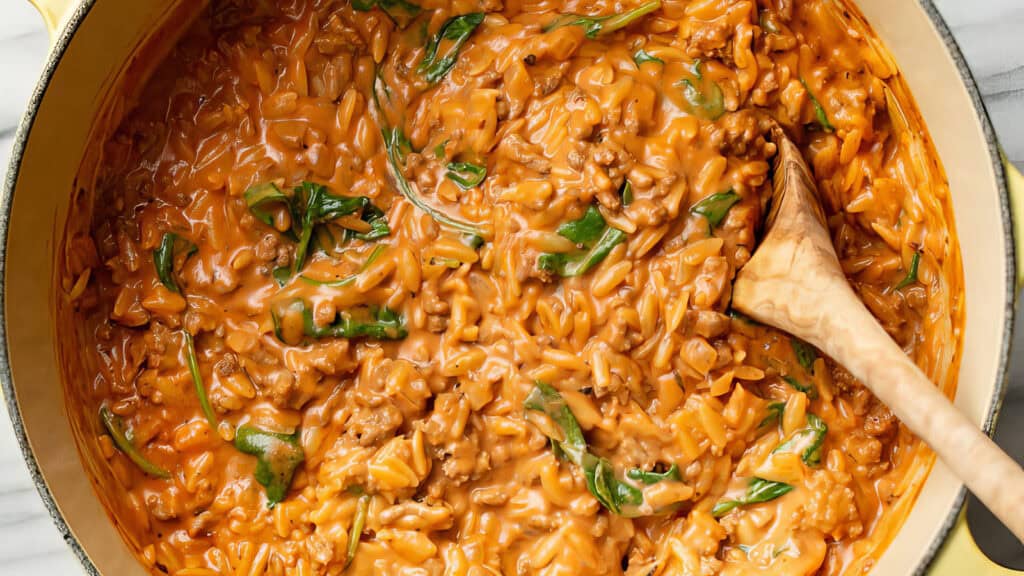 A pot filled with a creamy tomato-based pasta dish, cooked with ground beef and spinach. A wooden spoon is resting inside the pot, slightly stirring the mixture. The dish has a rich, orange sauce coating the pasta and other ingredients, making it one of the best ground beef recipes to try.