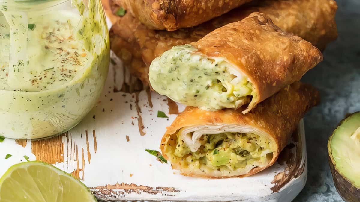 A close-up of golden-brown, crispy chimichangas filled with creamy avocado and greens, partially cut open to show the nourishing filling. They are served on a white rustic wooden board with a slice of lime and a jar of green dipping sauce beside them, highlighting both flavor and nutrition benefits.