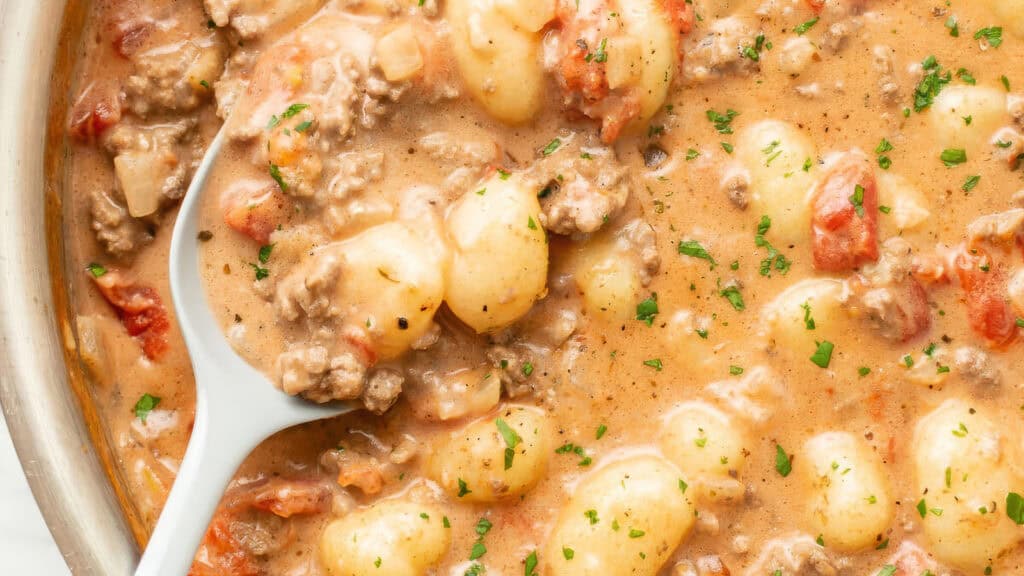 A close-up view of a creamy gnocchi dish, one of the best ground beef recipes, featuring tomatoes and herbs. The dish is in a pan and being stirred with a white spoon. Chopped parsley is sprinkled on top as garnish.