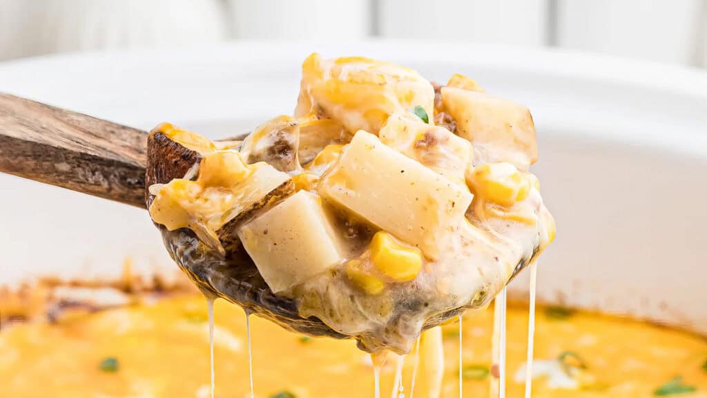 A wooden spoon holds a serving of cheesy potato casserole, featuring tender potato chunks, bits of corn, and melted cheese, with strings of cheese pulling as the spoon is lifted. Ideal for slow cooker recipes, the background shows the edge of a white casserole dish.