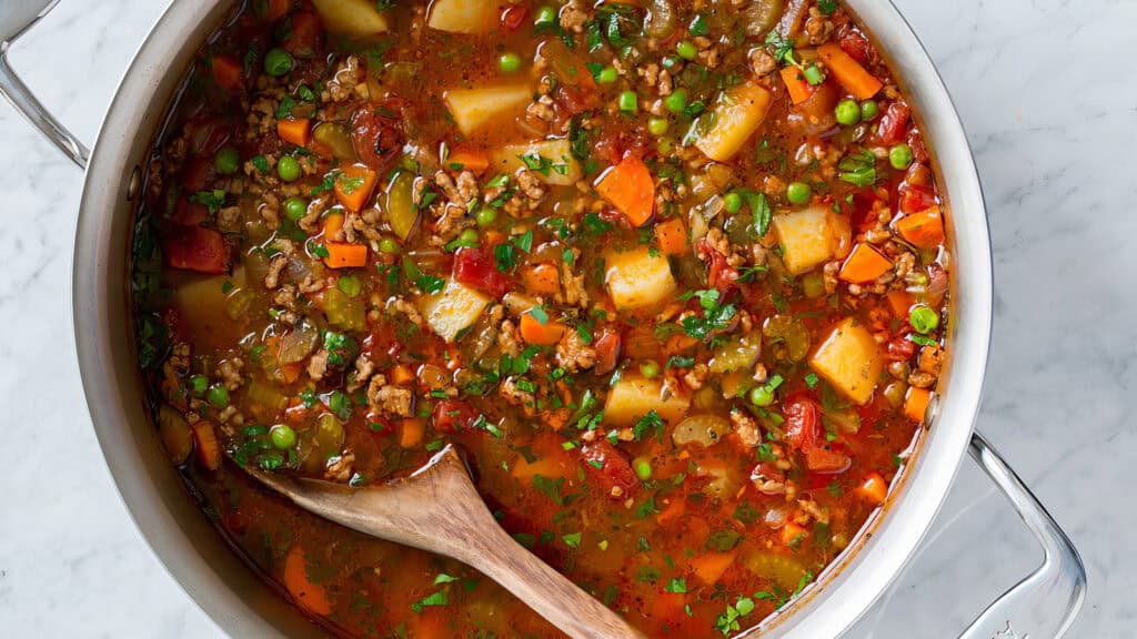 A metal pot filled with hearty vegetable soup. The colorful mixture includes diced carrots, potatoes, red peppers, peas, and tomatoes in a vibrant broth. Perfect for ground beef recipes, a wooden spoon rests inside the pot, ready for serving. The pot sits on a marble countertop.