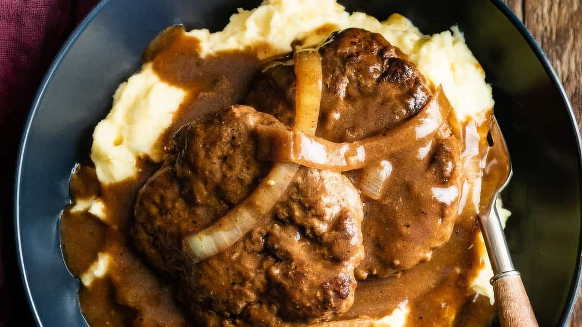 A plate of mashed potatoes topped with two Salisbury steaks covered in brown gravy and caramelized onion slices. A fork rests at the side of the black plate. The dish, inspired by comfort food recipes, is garnished with sauce, suggesting a hearty and comforting meal.