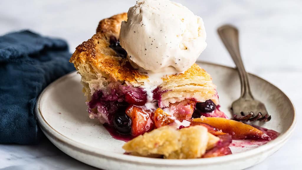 A slice of mixed berry and peach pie with a flaky crust, topped with a scoop of vanilla ice cream, served on a white plate with a fork. A blue cloth napkin is in the background. The juicy and vibrant pie filling contrasts beautifully with the golden-brown crust—a perfect treat among summer desserts.