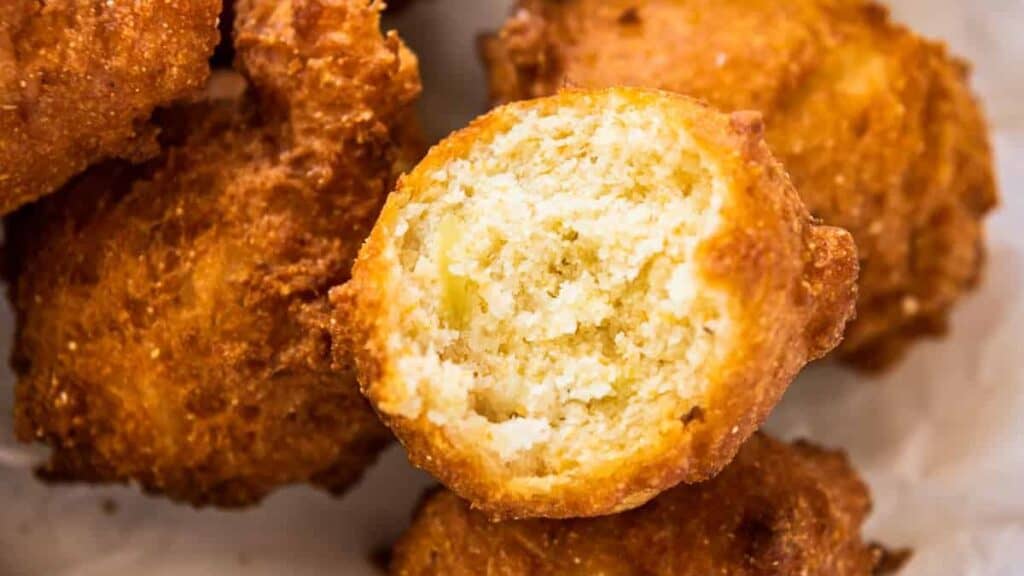 Close-up of golden brown hush puppies, with one showing a bite taken out, revealing a soft, crumbly inside. The hush puppies are slightly irregular in shape, highlighting their crispy, fried exterior—a classic touch in Southern recipes.