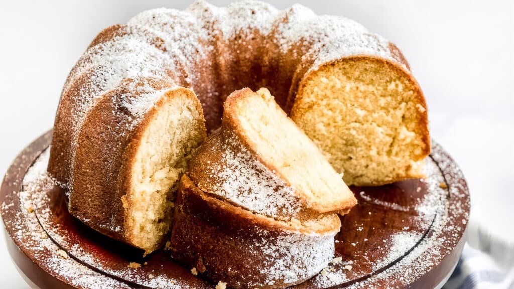 A Bundt cake dusted with powdered sugar sits on a round wooden platter. Several slices have been cut from the cake, revealing its moist and fluffy interior. The backdrop is a simple, light-colored surface that emphasizes the cake as the focal point, perfect for those seeking delightful Bundt Cake recipes.