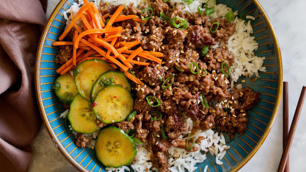 A colorful dish featuring white rice topped with seasoned ground beef, sliced carrots, and cucumber slices arranged neatly in a blue bowl. Chopped green onions and sesame seeds garnish the meal. A napkin and chopsticks are placed beside the bowl—one of our favorite ground beef recipes.