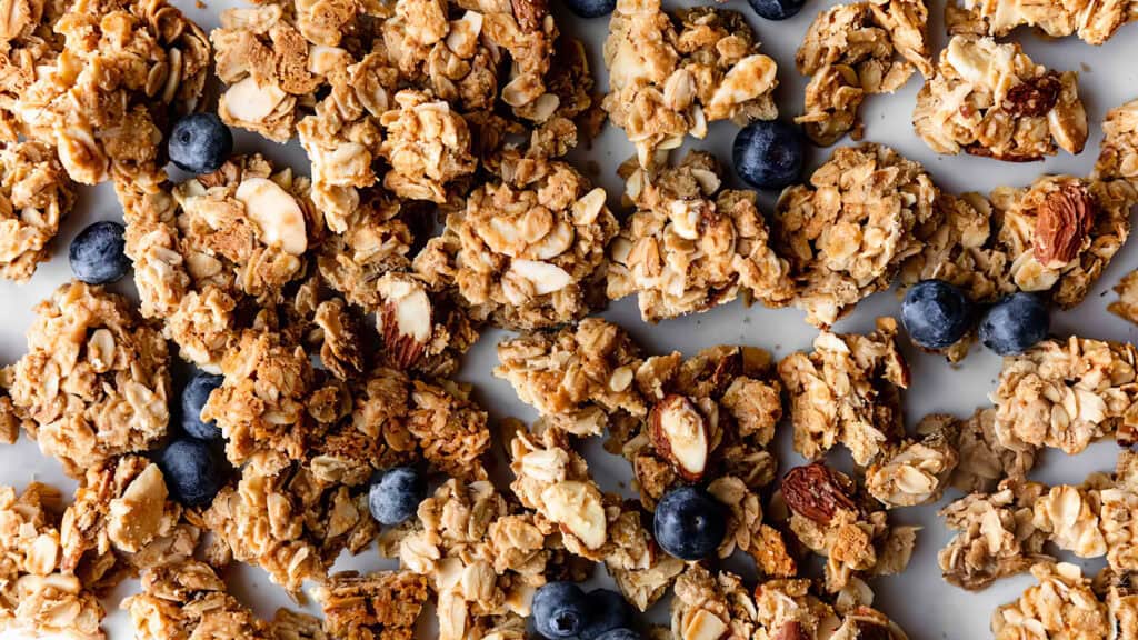 Close-up of clusters of granola mixed with sliced almonds, oats, and fresh blueberries spread out on a white surface. The granola appears crunchy and golden-brown, while the blueberries add a pop of vibrant blue color—perfect for your snack recipes.