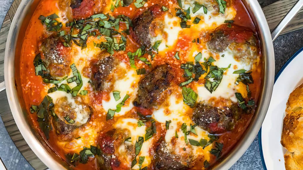 A close-up shot from "Meatball Magic" shows a skillet containing meatballs topped with melted cheese and garnished with chopped fresh basil leaves. The meatballs are covered in rich tomato sauce, and a piece of golden-brown bread can be seen on the side of the plate—perfect for cooking book inspiration.