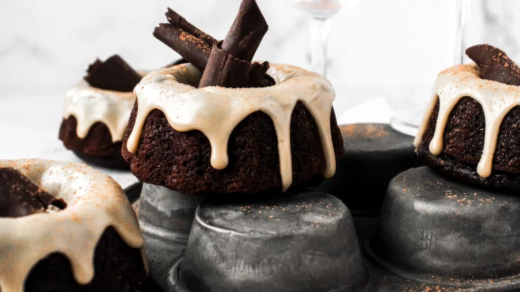 Close-up of chocolate bundt cakes with white icing dripping down the sides, each garnished with chocolate shards on top. Displayed on an upside-down muffin tin, these delectable treats are perfect for fans of Bundt Cake Recipes. The background is a blurred, neutral gray and white.