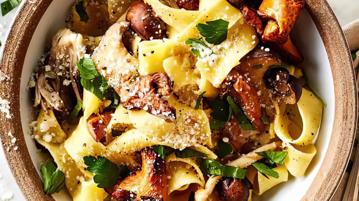 A close-up of a bowl of delicious pasta adorned with wild mushrooms; an easy, back-to-school recipe. The wide, flat noodles are mixed with sautéed mushrooms and garnished with fresh parsley and grated cheese. The dish looks fresh and inviting, capturing the textures and colors beautifully.