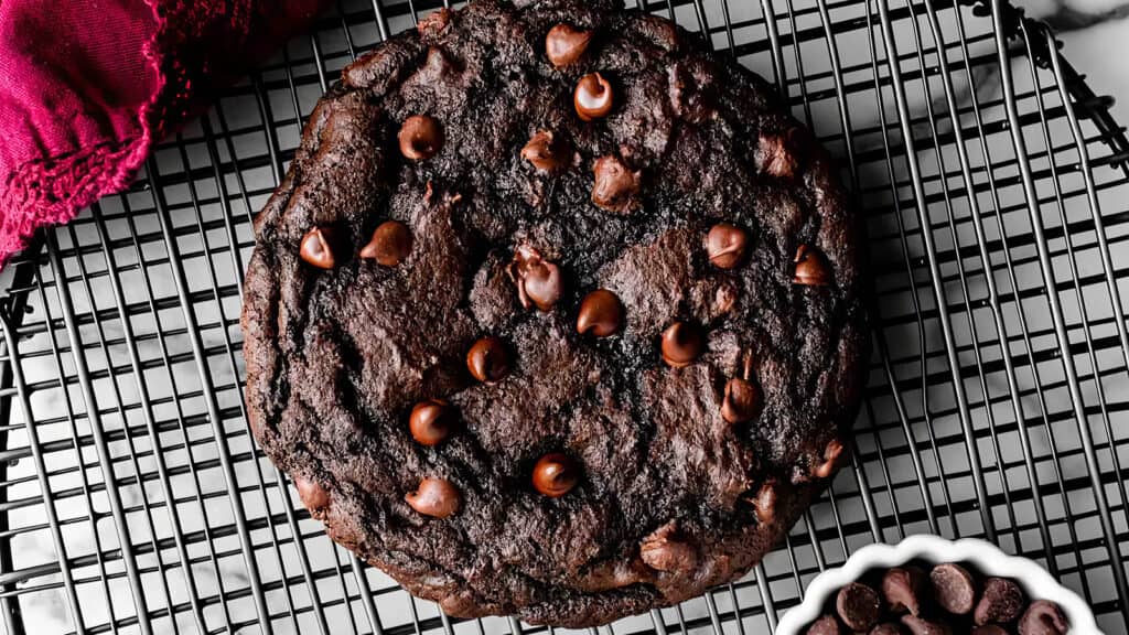 A large, freshly-baked chocolate chip cookie sits on a cooling rack, perfect for any chocolate recipe enthusiast. The cookie is dark brown with visible chocolate chips scattered on top. A red cloth is in the top left corner, and a small bowl of chocolate chips is in the bottom right corner.