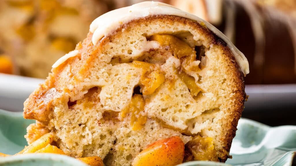 A close-up of a slice of decadent, moist peach bundt cake, showcasing its dense texture and chunks of fresh peaches throughout. The cake is topped with a light drizzle of white icing and served on a green plate with pieces of spiced peaches—perfect for those seeking bundt cake recipes.