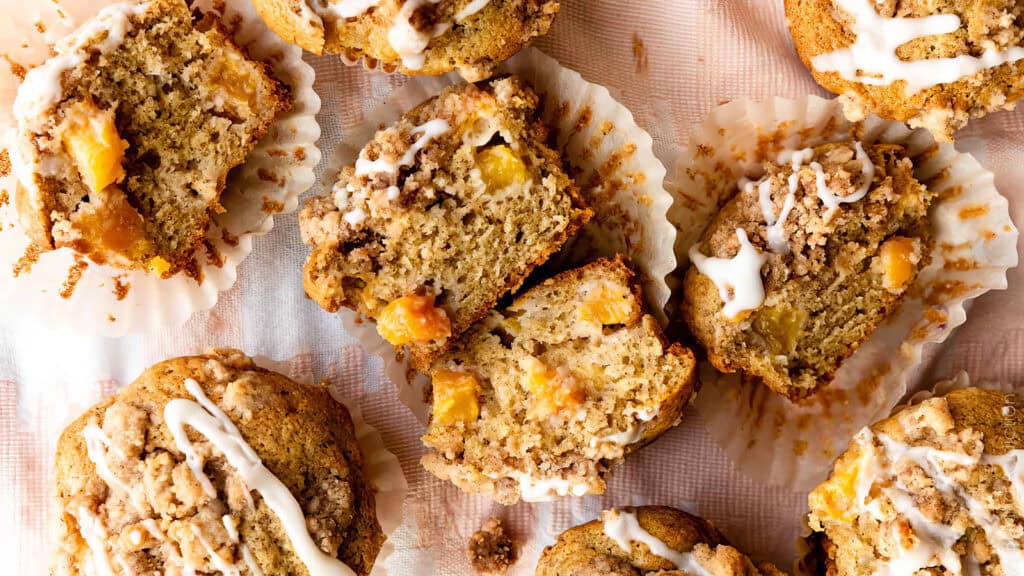 An overhead view of several peach muffins topped with icing. Some muffins are halved to show the peach chunks and moist interior. The muffins, exemplifying delicious muffin recipes, are arranged on crinkled parchment paper, creating a cozy and appetizing scene.