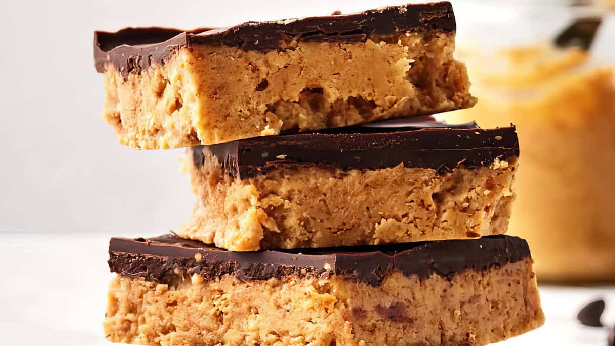 A close-up of three stacked chocolate peanut butter bars on a white surface. The bars have a thick peanut butter base and a smooth, glossy chocolate layer on top, perfect for satisfying cravings. A jar of peanut butter is slightly blurred in the background.