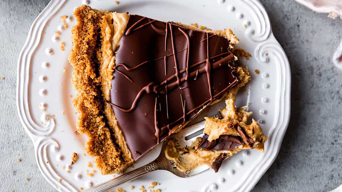 A slice of chocolate pie with a graham cracker crust is served on a white plate. Topped with smooth peanut butter and chocolate, a small piece has been cut off and is on a fork next to the slice. The plate features a delicate, embossed pattern around the edge, perfect for satisfying cravings.