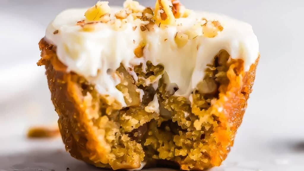 A close-up of a partially eaten carrot cake muffin topped with creamy white frosting and sprinkled with chopped nuts. The moist and fluffy texture of the muffin with visible carrot shreds is shown, with the light and smooth frosting melting slightly—perfect for those exploring new Muffin Recipes.