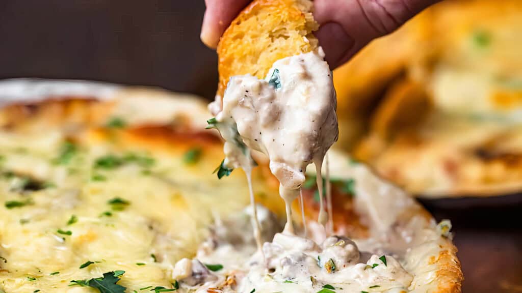 A hand dips a piece of bread into a creamy, gooey cheese and mushroom dip. The dip is in a shallow dish, topped with melted cheese and fresh herbs. This close-up scene showcases the rich texture and appetizing appearance, perfect for those seeking new dip recipes to try at home.