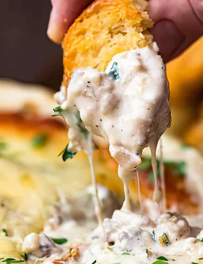 A hand dips a piece of bread into a creamy, gooey cheese and mushroom dip. The dip is in a shallow dish, topped with melted cheese and fresh herbs. This close-up scene showcases the rich texture and appetizing appearance, perfect for those seeking new dip recipes to try at home.