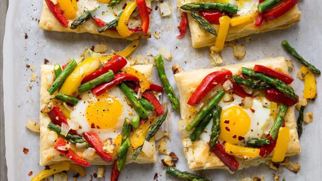 Four rectangular puff pastries topped with sunny-side-up eggs, red and yellow bell peppers, asparagus spears, and seasonings on parchment paper. The vibrant vegetables and eggs create a colorful and appetizing presentation reminiscent of the best pancake recipes.