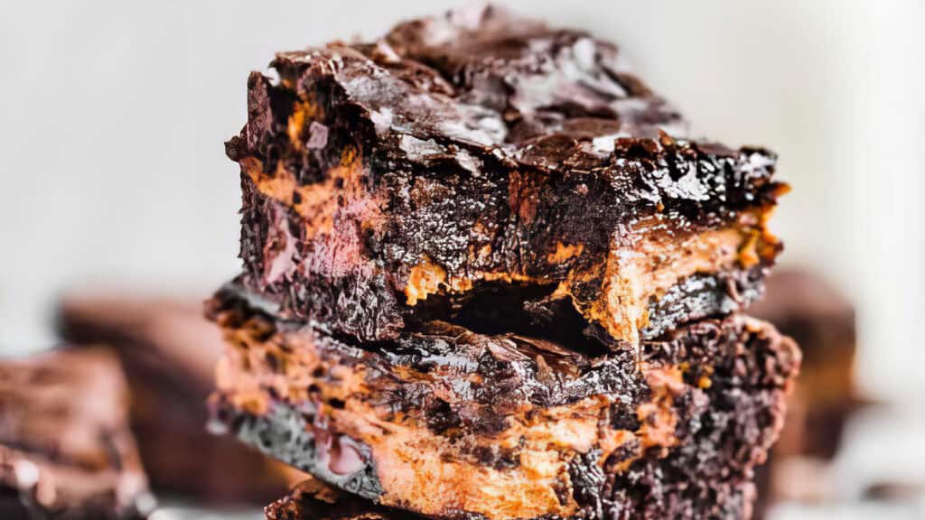 Close-up of a stack of two decadent brownies with a rich, gooey chocolate texture. Perfect for chocoholics, these brownies are layered with a ribbon of additional chocolate or fudge, highlighting their moist and indulgent interior. The background is blurred, emphasizing the beyond basic dessert.