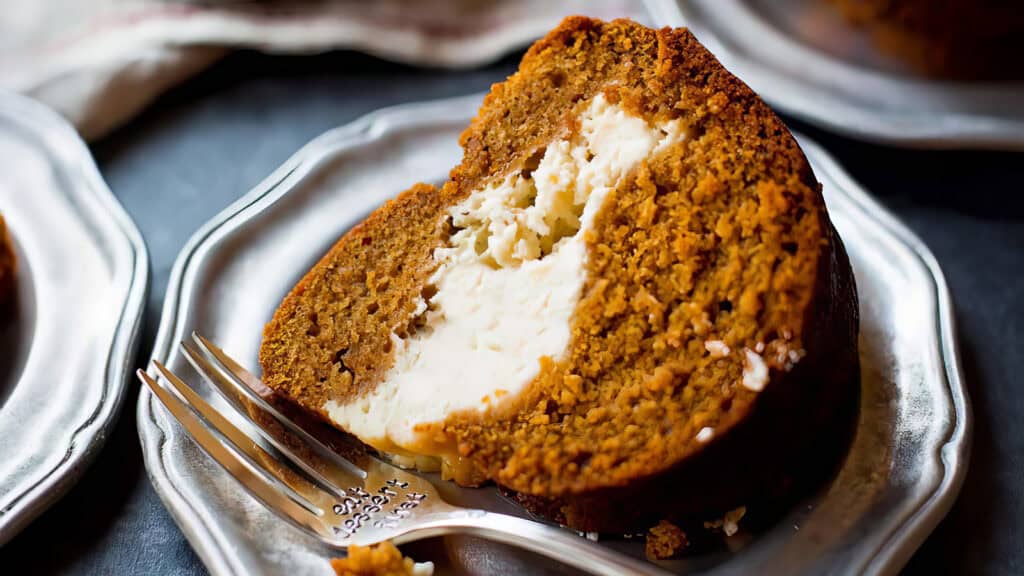 A close-up of a slice of pumpkin cake with a creamy filling on a silver plate. A fork rests on the plate beside the cake slice, showing a few crumbs near the cut piece. In the background, blurred portions of more cake slices on similar plates hint at irresistible Bundt Cake recipes.