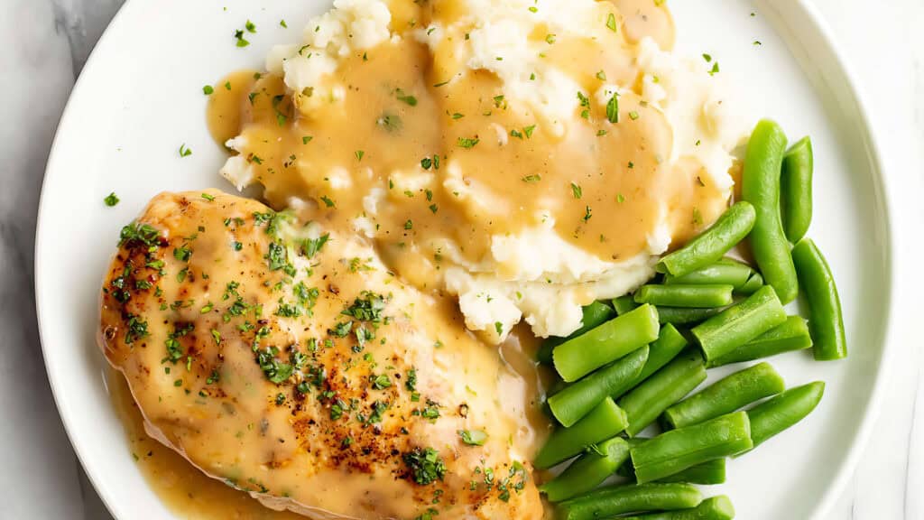 A white plate with a serving of mashed potatoes topped with gravy, a seasoned and herb-coated chicken breast with gravy, and a side of green beans, inspired by delightful chicken recipes. The meal is garnished with chopped parsley.