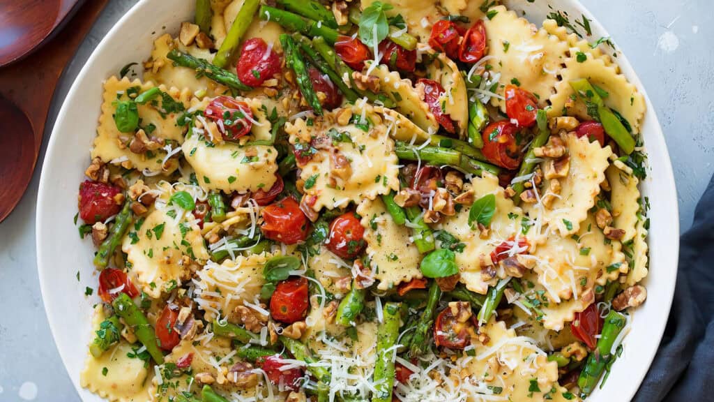 A close-up of a fresh ravioli pasta dish mixed with asparagus, cherry tomatoes, walnuts, and grated cheese garnished with parsley and basil. The colorful ingredients are spread evenly in a large white bowl, making it a delightful treat for Pasta Lovers.