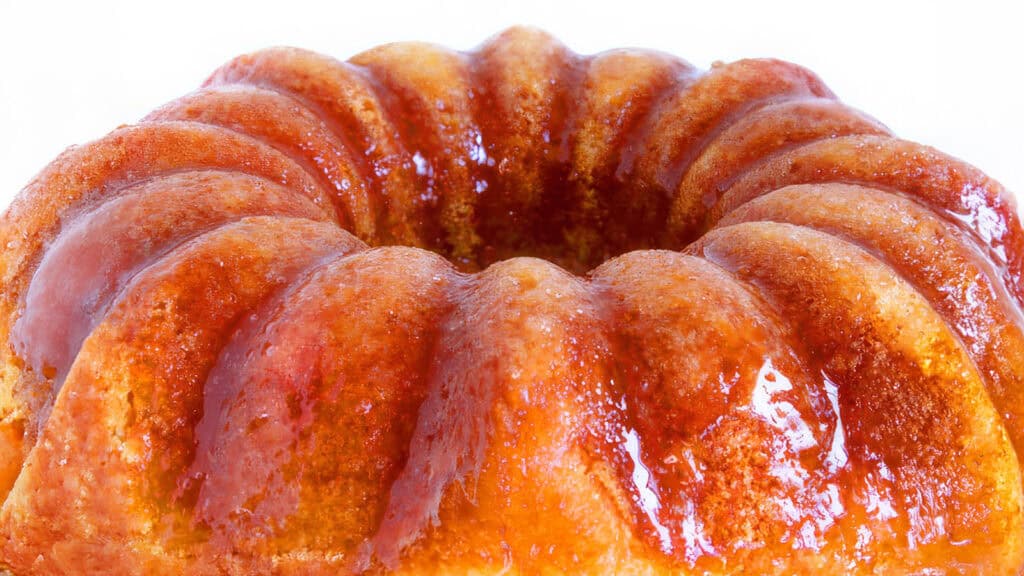 A close-up view of a glazed bundt cake with a golden-brown color and a shiny, glossy surface. The cake has a circular shape with fluted edges and looks moist and delicious, with the glaze enhancing its texture. Perfect for those exploring Bundt Cake Recipes, the background is white.