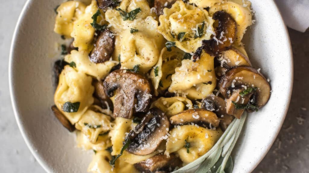 A white bowl filled with creamy tortellini pasta mixed with sautéed mushrooms and garnished with grated Parmesan cheese and chopped herbs. 