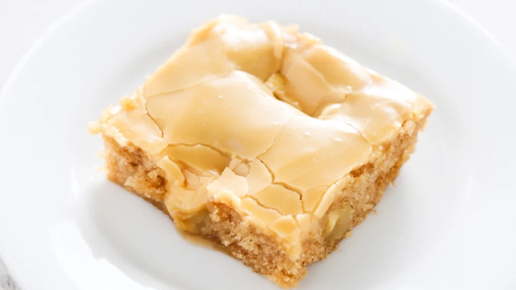 A close-up of a square piece of apple cake with a glossy, beige-colored glaze on top, served on a white plate. The cake appears moist with visible apple chunks and a cracked surface glaze, perfect for those looking to try new Sheet Cake Recipes.