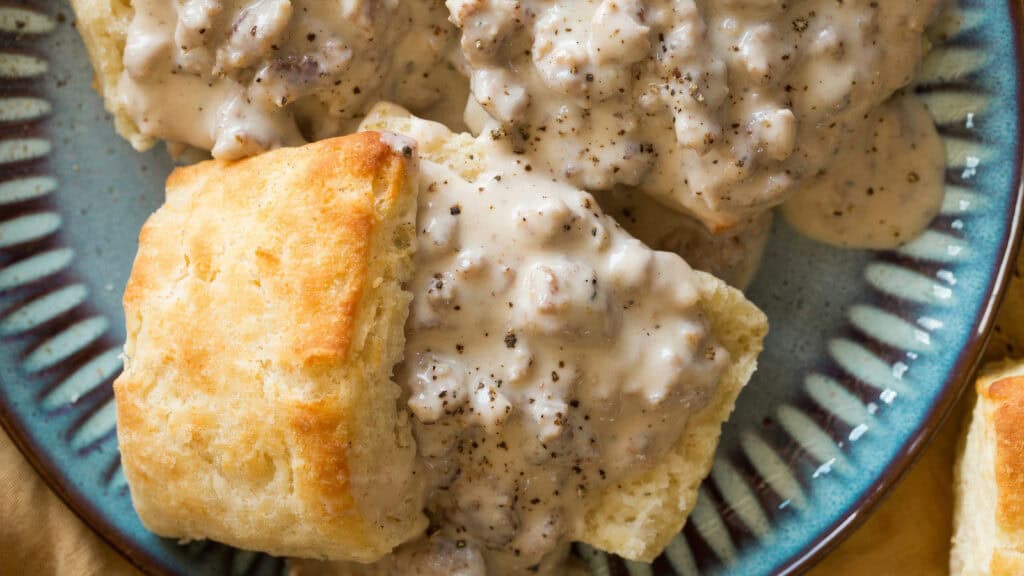 A plate of golden-brown biscuits covered in creamy sausage gravy, garnished with black pepper. The flaky biscuits and rich gravy with chunks of sausage embody the essence of Southern recipes, presented on a blue dish with a detailed rim.