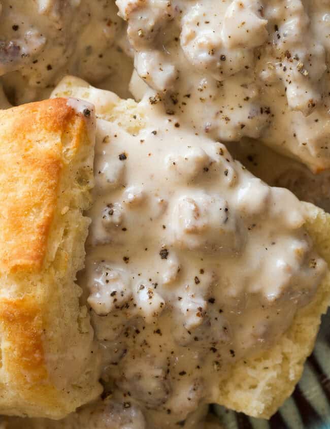 A plate of golden-brown biscuits covered in creamy sausage gravy, garnished with black pepper. The flaky biscuits and rich gravy with chunks of sausage embody the essence of Southern recipes, presented on a blue dish with a detailed rim.