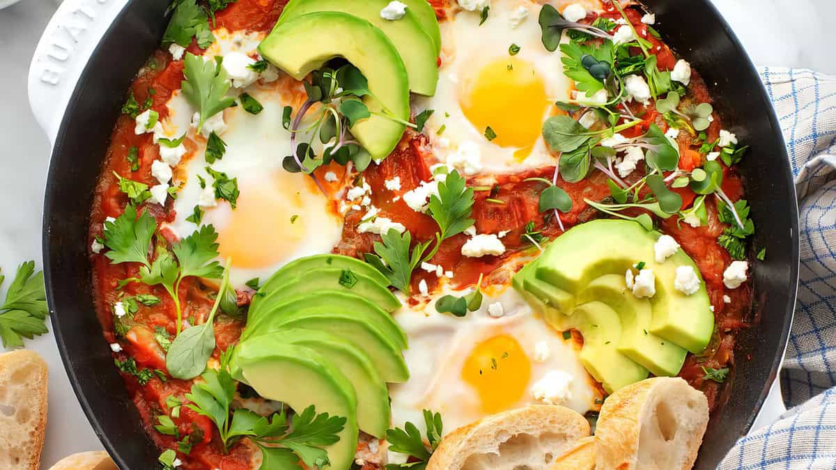 A skillet filled with shakshuka: poached eggs in a spiced tomato sauce, garnished with sliced avocado, crumbled feta, and fresh greens. Slices of crusty bread are placed nearby, ready for dipping. This vibrant and appetizing dish is perfect for Lunch and Dinner or even as an easy egg recipe for Breakfast.