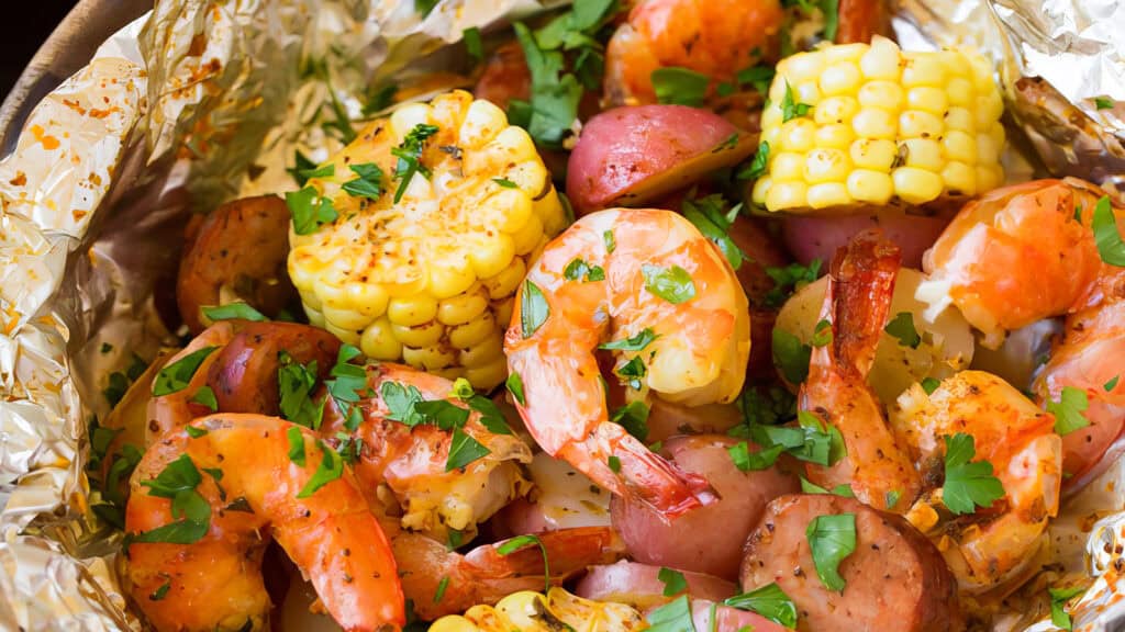 A foil packet filled with a shrimp boil, featuring plump shrimp, savory sausage slices, chunks of corn on the cob, and pieces of red potato. The contents are garnished with chopped parsley, bringing a touch of Southern recipes to your meal.