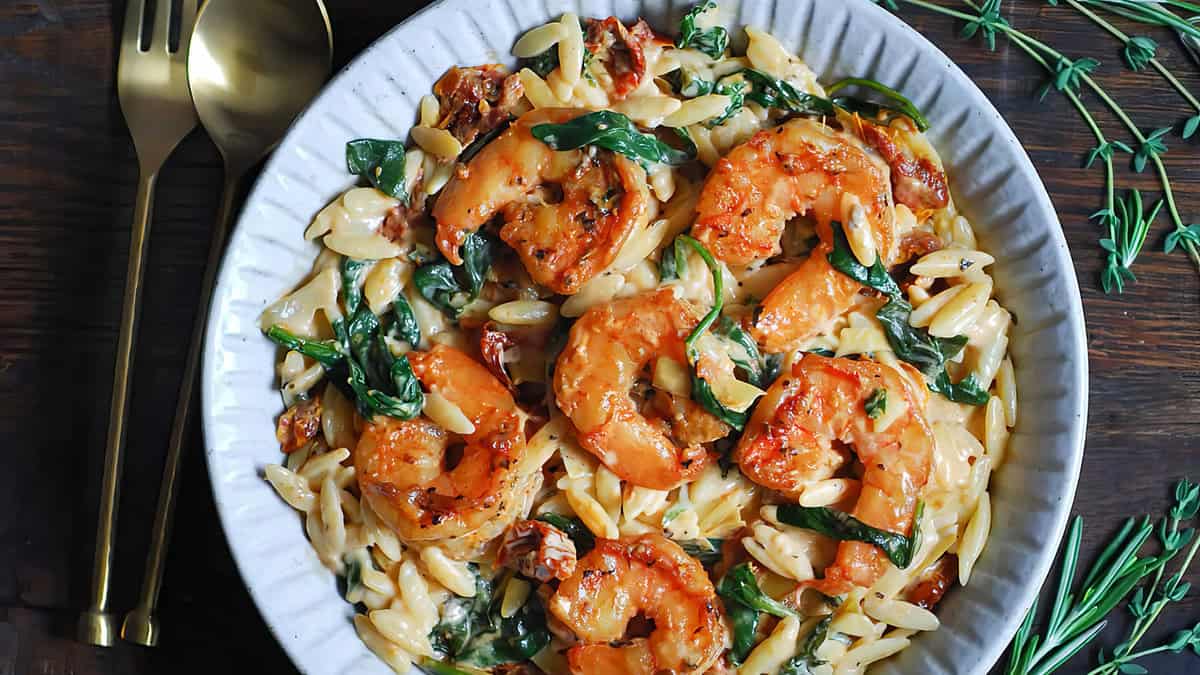 A white bowl filled with creamy orzo pasta topped with sautéed shrimp, sun-dried tomatoes, and spinach. The dish is garnished with fresh herbs and served with a gold fork and spoon on a wooden table—perfect for Back-to-School recipes that are easy peasy.