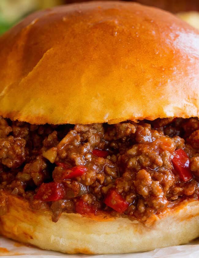 A close-up of a sloppy joe sandwich on a shiny toasted bun, filled with a generous serving of seasoned ground beef and diced red peppers—perfect for anyone exploring ground beef recipes. The sandwich is accompanied by side items, including fresh greens, potato chips, and pickle slices.