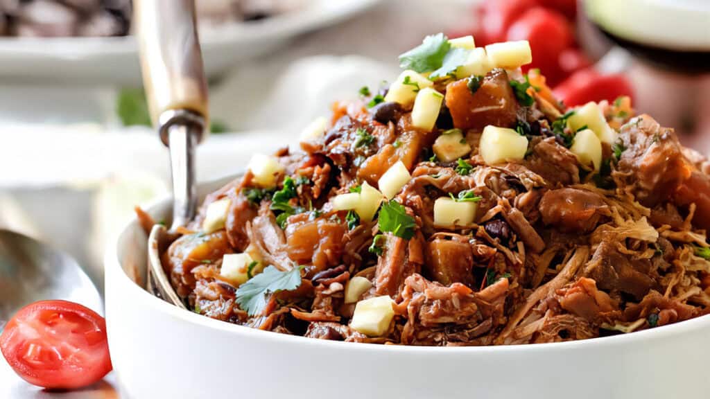 A white bowl filled with tender pulled pork from our favorite slow cooker recipes, topped with diced cheese and garnished with fresh cilantro. There are slices of tomatoes and a fork resting on the edge of the bowl. The background is blurred, showing more food items.