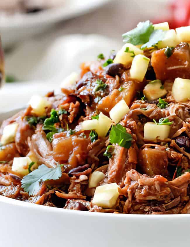 A white bowl filled with tender pulled pork from our favorite slow cooker recipes, topped with diced cheese and garnished with fresh cilantro. There are slices of tomatoes and a fork resting on the edge of the bowl. The background is blurred, showing more food items.