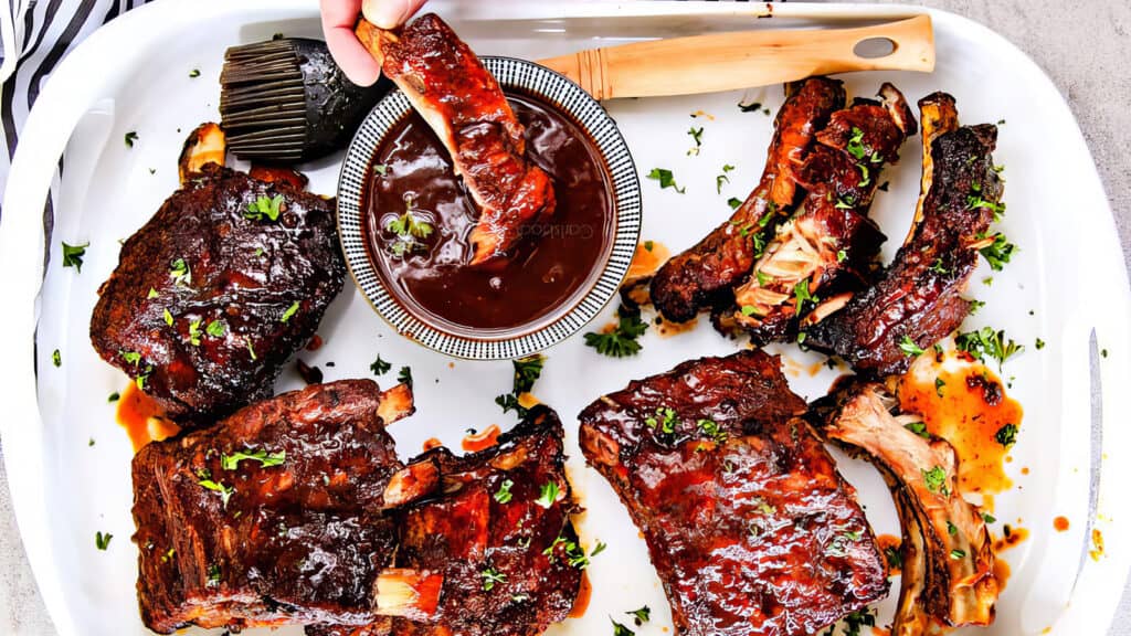 A white tray holds several portions of barbecued ribs garnished with chopped herbs. One rib is being dipped into a bowl of barbecue sauce in the center of the tray, reminiscent of slow cooker recipes. A wooden-handled grill brush covered in sauce rests nearby.