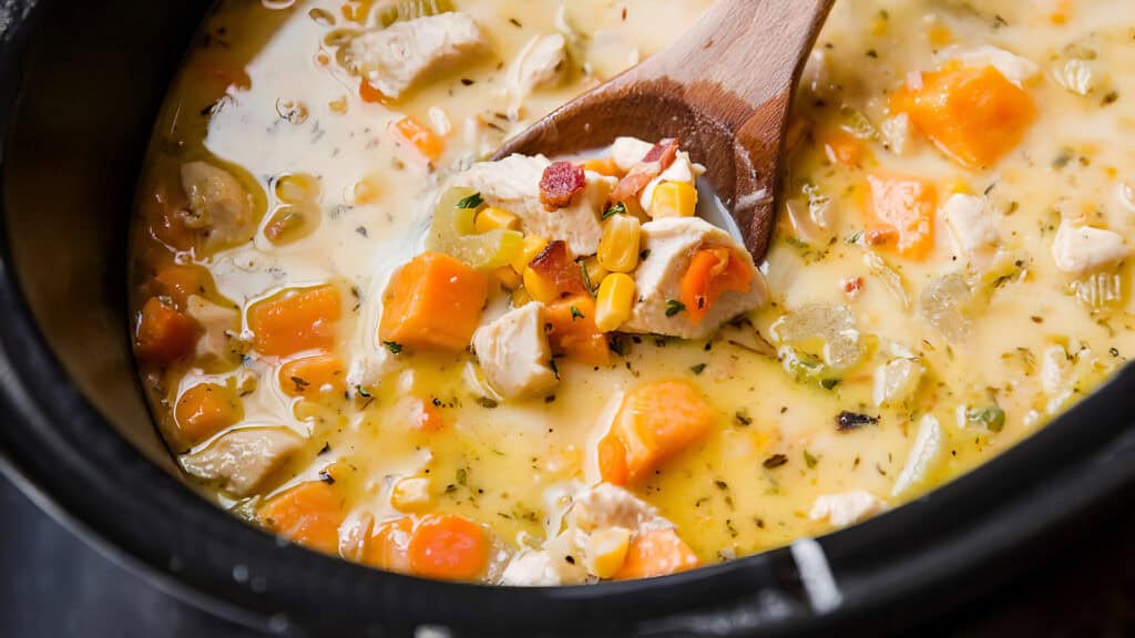 A close-up of a hearty chicken and vegetable soup in a black pot. The slow cooker recipe features chunks of chicken, corn, celery, carrots, and sweet potatoes in a creamy broth. A wooden spoon is partially submerged, lifting up several ingredients.