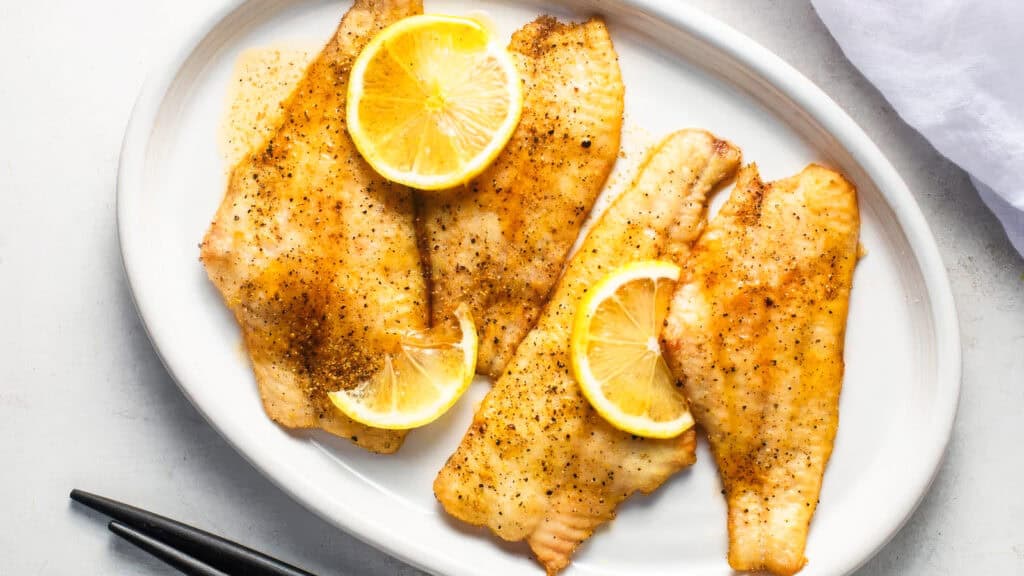 A white oval plate contains four pieces of seasoned and grilled white fish fillets garnished with lemon slices, reminiscent of Southern recipes. The plate is set on a light-colored surface, with chopsticks placed beside it. A white cloth is partially visible in the top right corner.