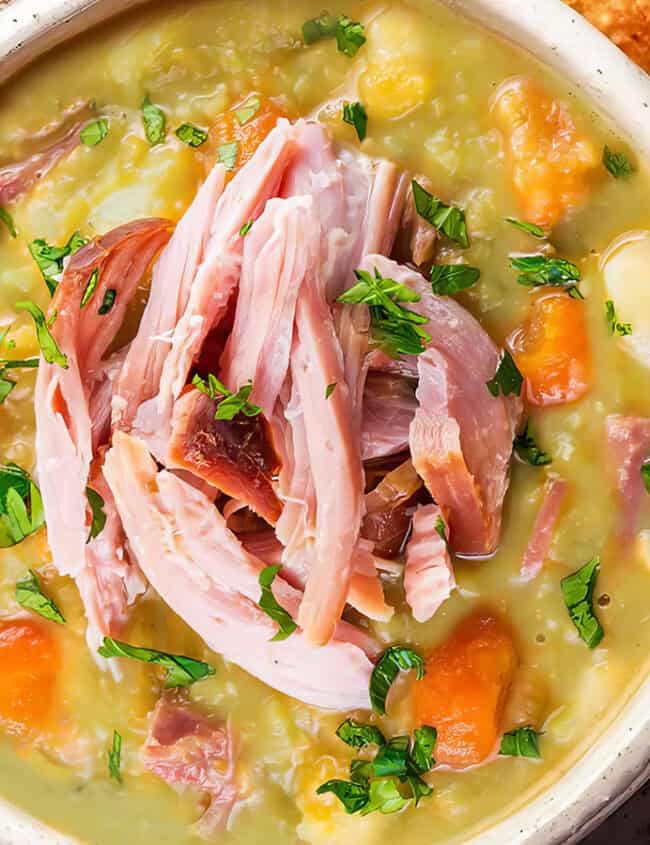 A bowl of hearty pea and ham soup featuring chunks of ham, diced carrots, and garnished with fresh parsley. The soup has a creamy consistency and a comforting appearance. Beside the bowl, slices of toasted bread are arranged on a ceramic plate—perfect for comfort food recipes.