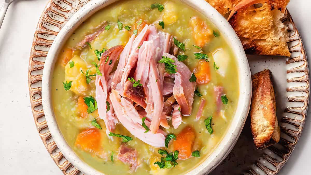 A bowl of hearty pea and ham soup featuring chunks of ham, diced carrots, and garnished with fresh parsley. The soup has a creamy consistency and a comforting appearance. Beside the bowl, slices of toasted bread are arranged on a ceramic plate—perfect for comfort food recipes.