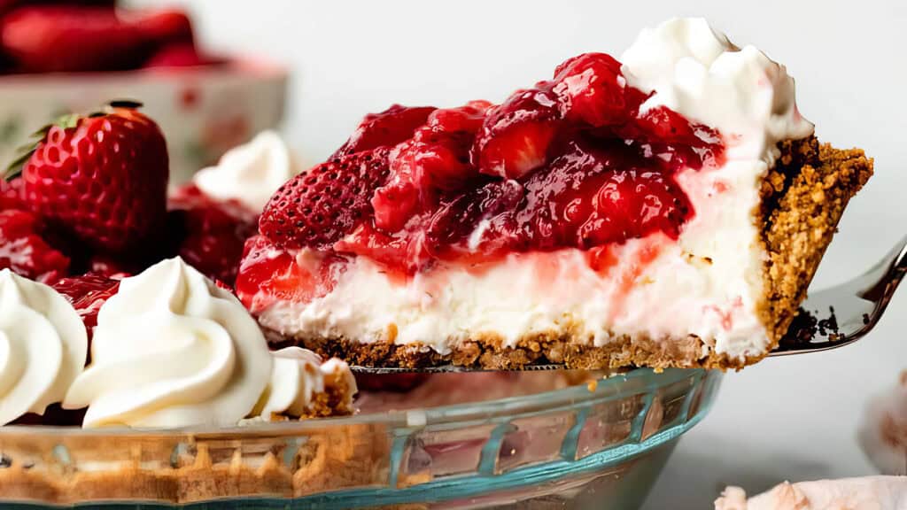 A close-up of a slice of strawberry cheesecake, one of the finest strawberry desserts, being lifted out of a glass pie dish. The cheesecake has a thick graham cracker crust, a creamy filling, and is topped with a generous layer of strawberry topping and fresh strawberries. Whipped cream adorns the edge.