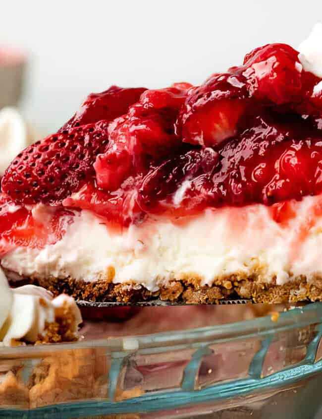 A close-up of a slice of strawberry cheesecake, one of the finest strawberry desserts, being lifted out of a glass pie dish. The cheesecake has a thick graham cracker crust, a creamy filling, and is topped with a generous layer of strawberry topping and fresh strawberries. Whipped cream adorns the edge.